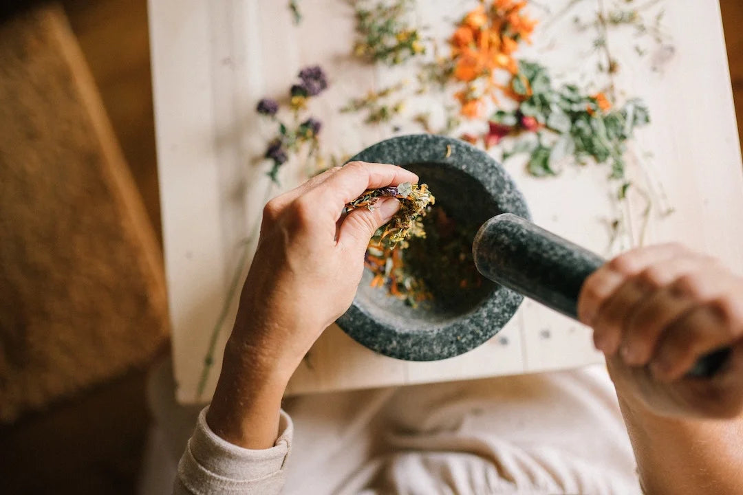 A person adding more herbs to a pestle and mortar, with dried flowers on the table 